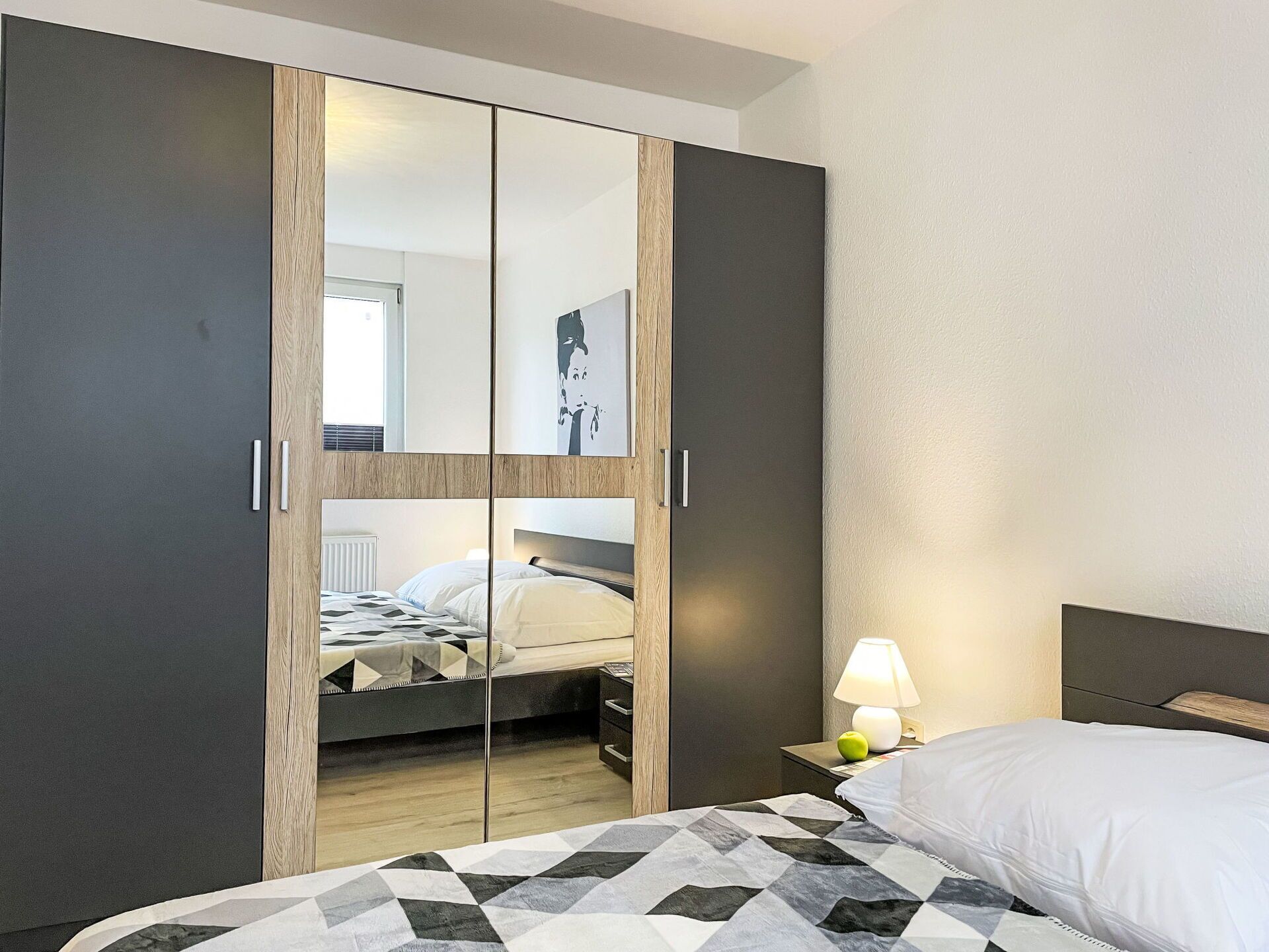 A bedroom with a bed, a nightstand with a lamp, and a wardrobe with a mirror.