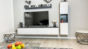 Uma sala de estar moderna com uma grande TV de tela plana presa a uma parede branca, uma mesa de centro de vidro com frutas e um tapete estampado.