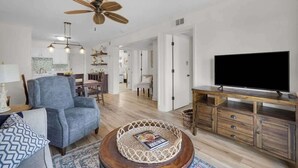 A modern living room with a blue armchair, a wooden coffee table, a TV on a wooden stand, and a ceiling fan. - Tennis Villa C14 - Easy Walk to the Beach! (Hilton Head Island)