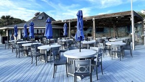 An outdoor dining area with round tables and chairs, covered by blue umbrellas, and a covered seating area in the background. - Tennis Villa C14 - Easy Walk to the Beach! (Hilton Head Island)