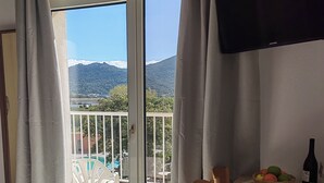 A room with a bed, a dining table with chairs, a TV mounted on the wall, and a view of a balcony with outdoor furniture and a mountainous landscape. - Ferienhotel Maristella  (Aregno)