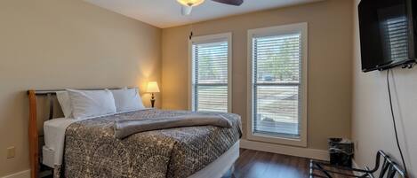 A bedroom with a bed, ceiling fan, two windows with blinds, a TV mounted on the wall, and a chair.