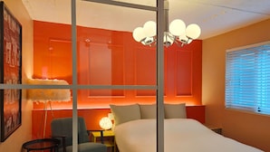 Une chambre d’hôtel avec un grand lit, une chaise, une petite table avec une lampe et une fenêtre avec des stores.