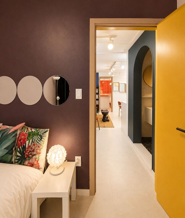 Un intérieur moderne comprenant un lit, une table de chevet, une lampe et un couloir menant à un espace cuisine.