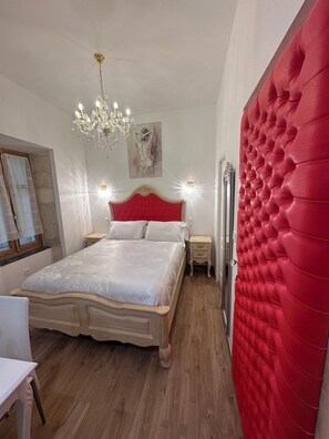 A bedroom with a large bed, a chandelier, a red padded wall, and a painting on the wall. - Small studio Presbytery in the Church (Narbonne)