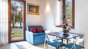 A room with a blue sofa, a dining table with chairs, a painting on the wall, and a window with a view of trees. - Cozy Beachfront House with Stunning Sea View | Mandriola - Putzu Idu (Mandriola)