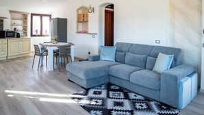 Ein modernes Wohnzimmer mit einem grauen Ecksofa, einem Esstisch mit Stühlen, einer Küchenzeile inklusive Mikrowelle und Schränken sowie einem gemusterten Teppich.