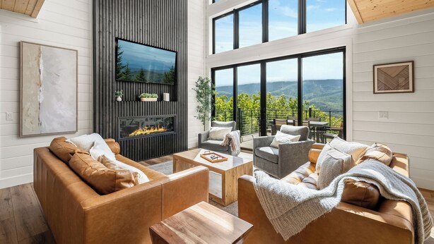 A spacious living area with leather sofas, a wooden coffee table, and a large TV mounted on a wall. There is a fireplace with a view of the outdoors, and large windows offering a scenic view of the mountains. - MtnTopRetreat: MtnView*Pool*Theater*Games*HotTub*WiFi*Fireplaces*ResortPool (Gatlinburg)