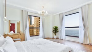 A bedroom with a large bed, a wooden dresser, a mirror, a chandelier, and a view of the sea through large windows. - Savis Home – Oceanfront Serenity & Marina Views (Dubai)