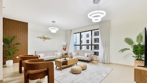 A modern living room with a sofa, two armchairs, a coffee table, and a TV stand. There are large windows with curtains, a potted plant, and a hanging light fixture. - Savis Home – Oceanfront Serenity & Marina Views (Dubai)