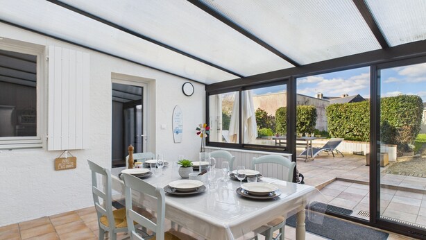 Een eetgedeelte met een tafel gedekt voor vier personen, witte stoelen, een klok aan de muur en uitzicht op de tuin door grote ramen.