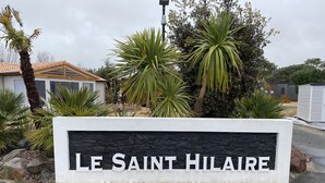 A sign reading "LE SAINT HILAIRE" with a stone facade and white lettering, surrounded by tropical plants and a residential area in the background. - Chalet for 6 in St-Hilaire - Heated swimming pool (Saint-Hilaire-de-Riez)