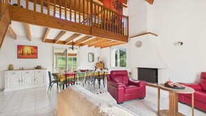 Ein geräumiger Wohnbereich mit hoher Decke, Holzbalken, einem Essbereich mit Tisch und Stühlen, einem roten Ledersofa und einem Kamin.