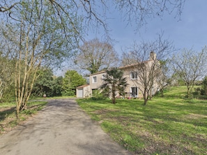 A two-story house with a paved driveway leading to the entrance, surrounded by trees and greenery.