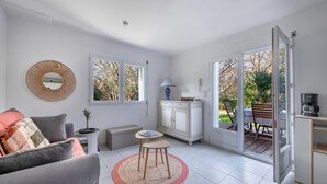 Ein modernes Wohnzimmer mit einer Couch, einem kleinen runden Tisch und einem Schrank. Zwei Fenster und eine Glastür führen in einen Außenbereich.