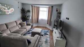 Ein Wohnzimmer mit beiger Couch, einem kleinen Tisch, einem Fernseher und einem Fenster mit Vorhängen.