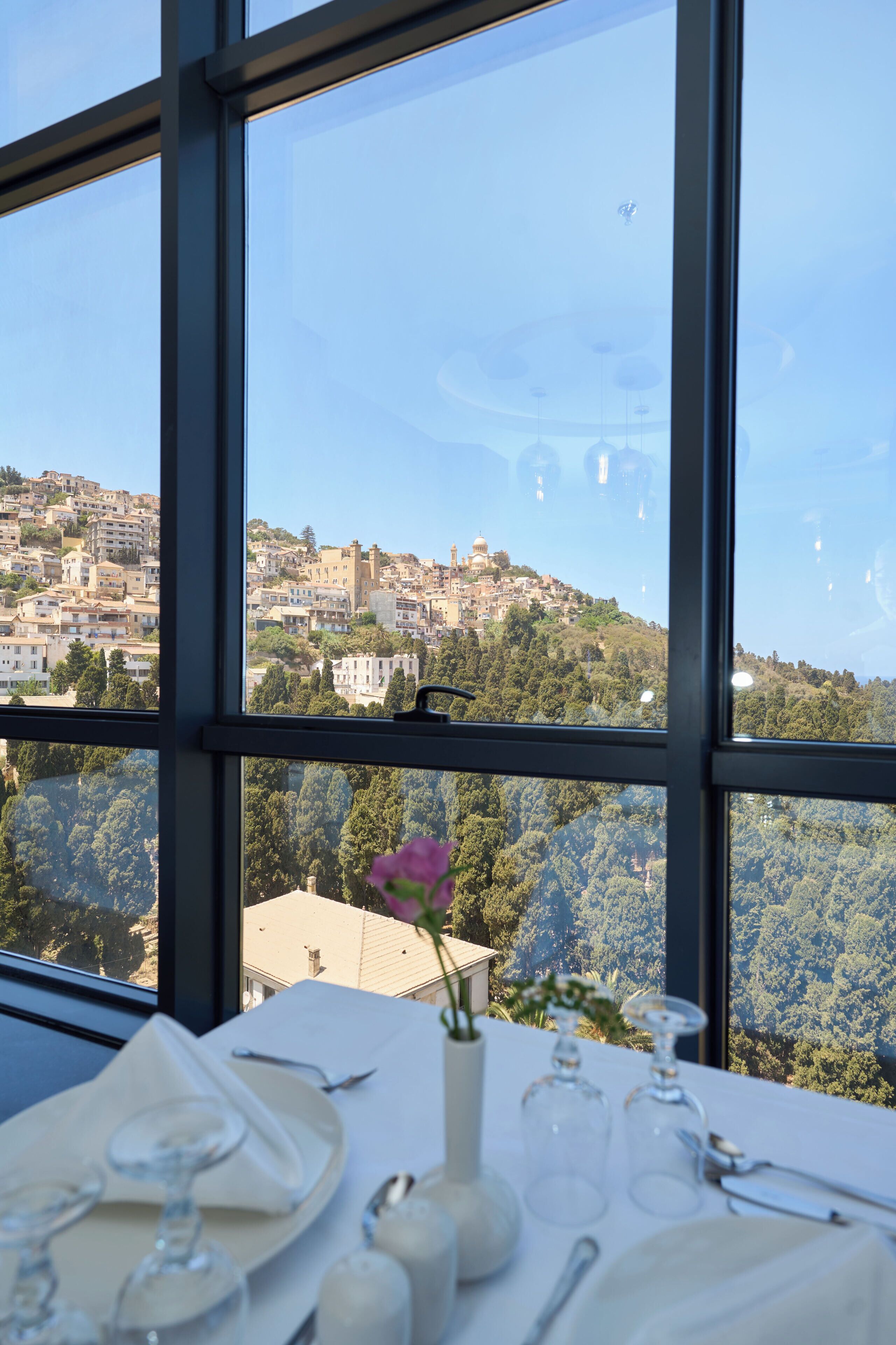 A table set for dining with a view of a hillside town through large windows.