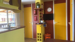 Una habitación con una mesa de madera, un armario amarillo, un estante con un reloj y un televisor colgado en la pared.