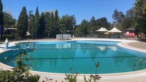 Piscina azul cristalina con un borde de baldosas, rodeada de árboles y zonas sombreadas.