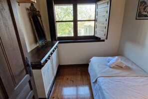 Une petite chambre avec un plancher en bois, un lit avec une literie blanche, une fenêtre avec des stores et une commode.