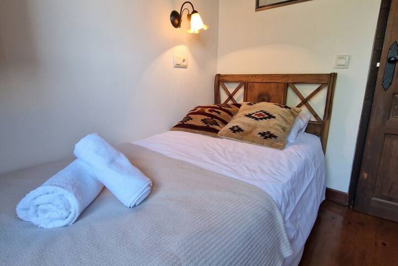 A single bed with a wooden headboard, a white blanket, two rolled towels, and patterned pillows.