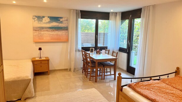 A bedroom with a bed, a bedside table, a nightstand, a window with curtains, a dining table with chairs, and a painting on the wall. - Beautiful apartment with garden seating area in Rapperswil-Jona (Rapperswil-Jona)