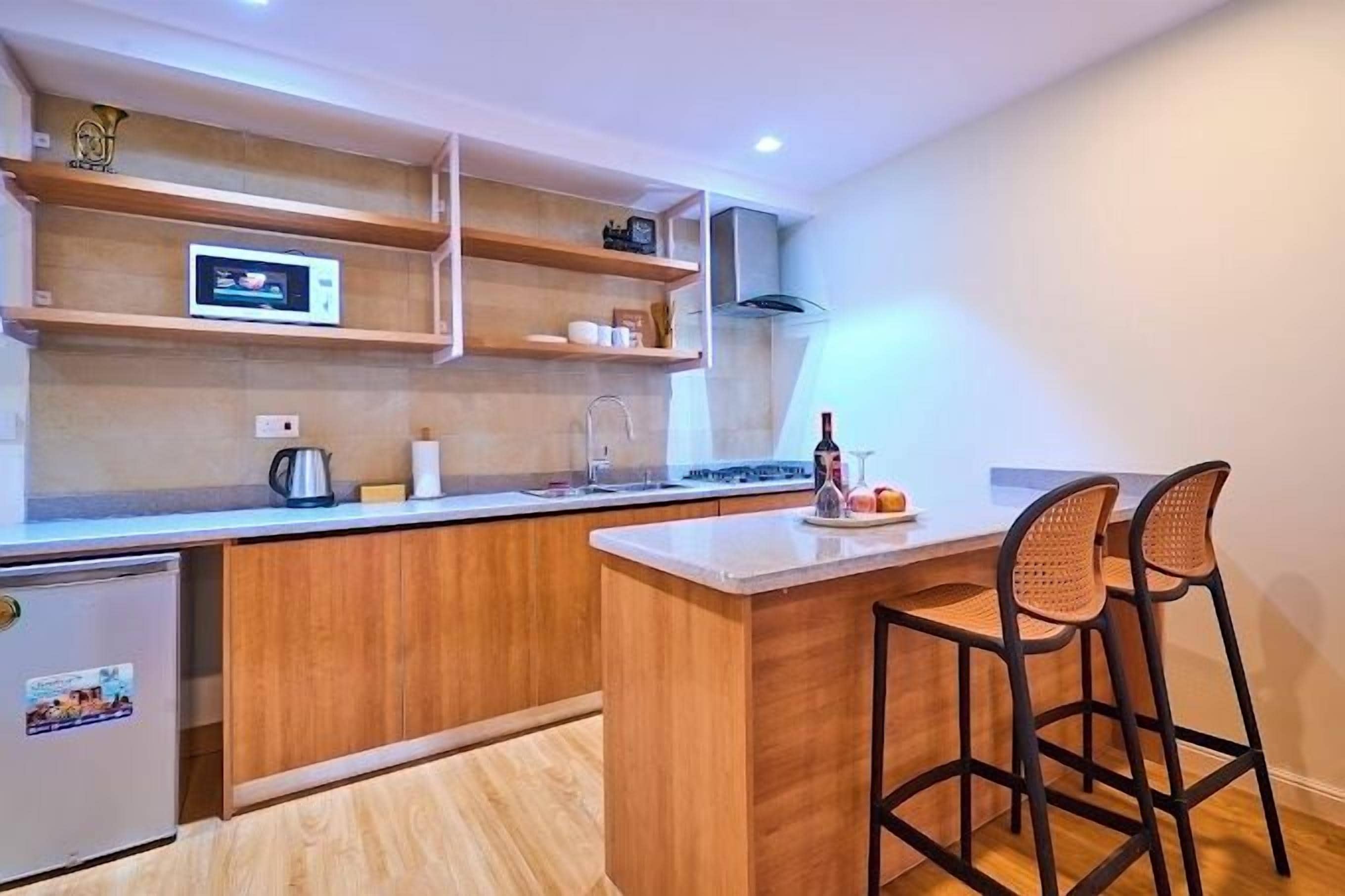 A compact kitchen with wooden cabinets, a built-in microwave, and a small refrigerator. There are two bar stools and a wine bottle on the counter.