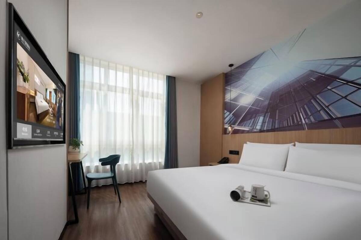 Habitación de hotel con una cama grande, un escritorio con silla, un televisor y un amplio ventanal con cortinas.