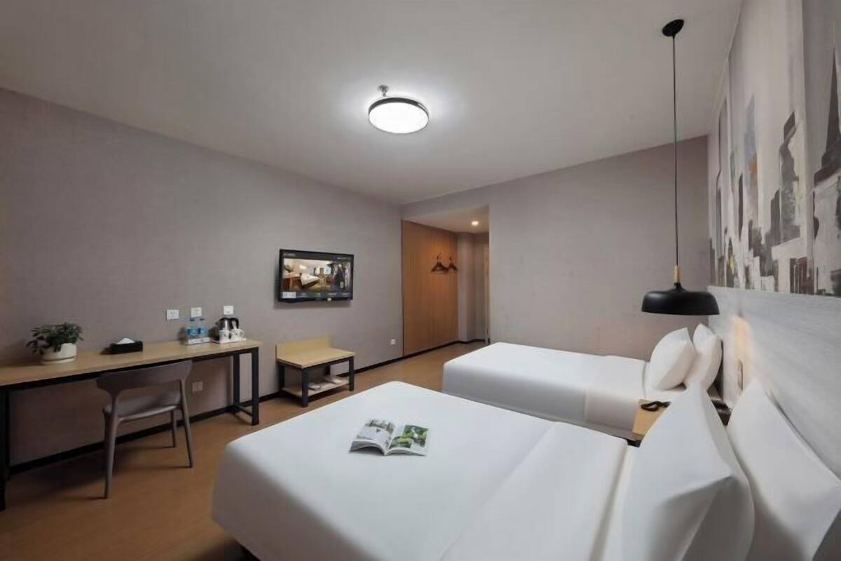 Habitación de hotel con dos camas, un escritorio, una silla, una mesita, una lámpara y un cuadro en la pared.