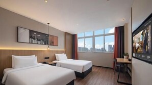 Une chambre d’hôtel avec deux lits, un bureau, une chaise, une fenêtre donnant sur la ville et un tableau encadré au mur.
