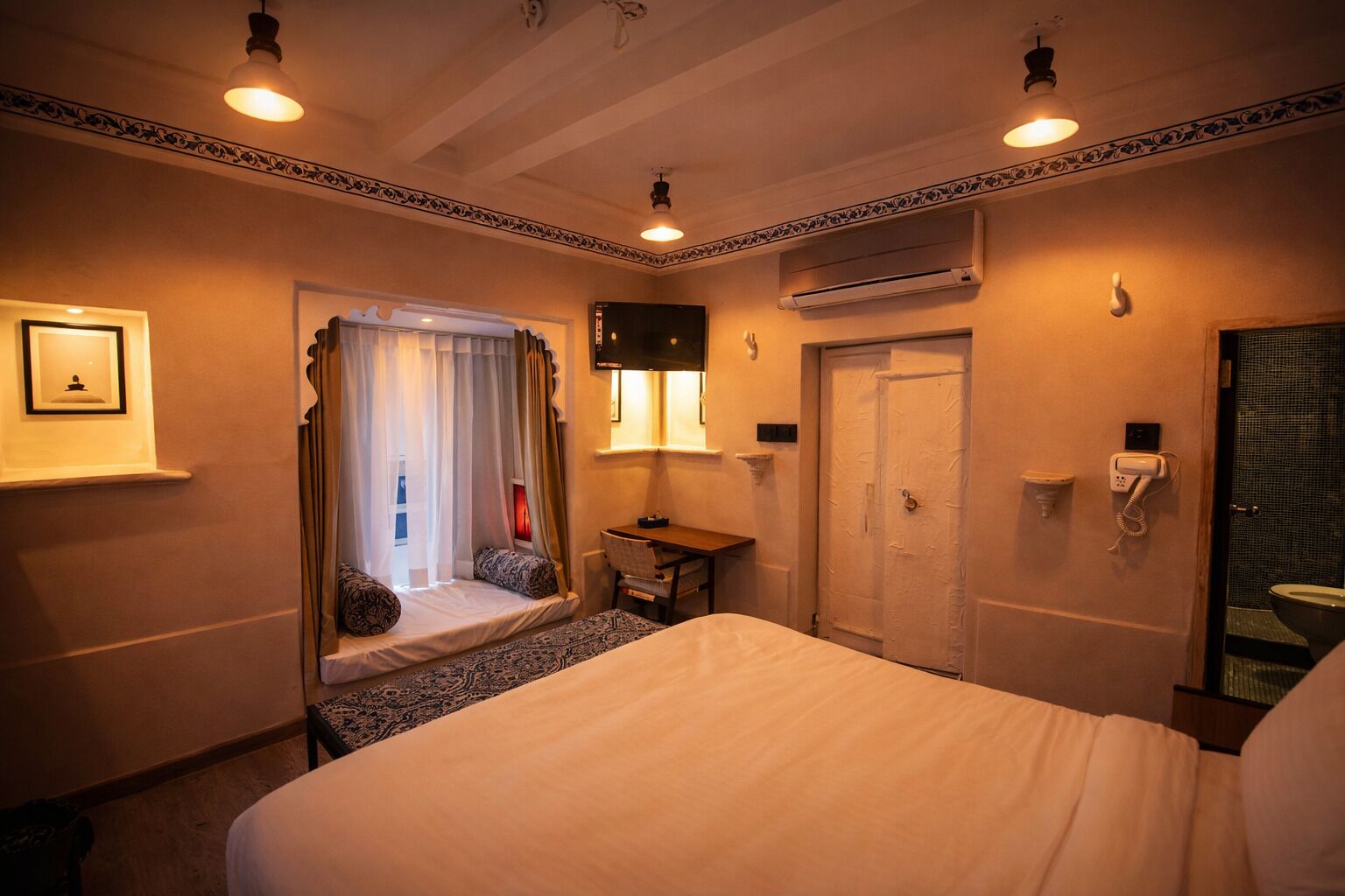 A hotel room with a bed, a desk, a chair, a window with curtains, an air conditioning unit, a telephone, and a framed picture on the wall.