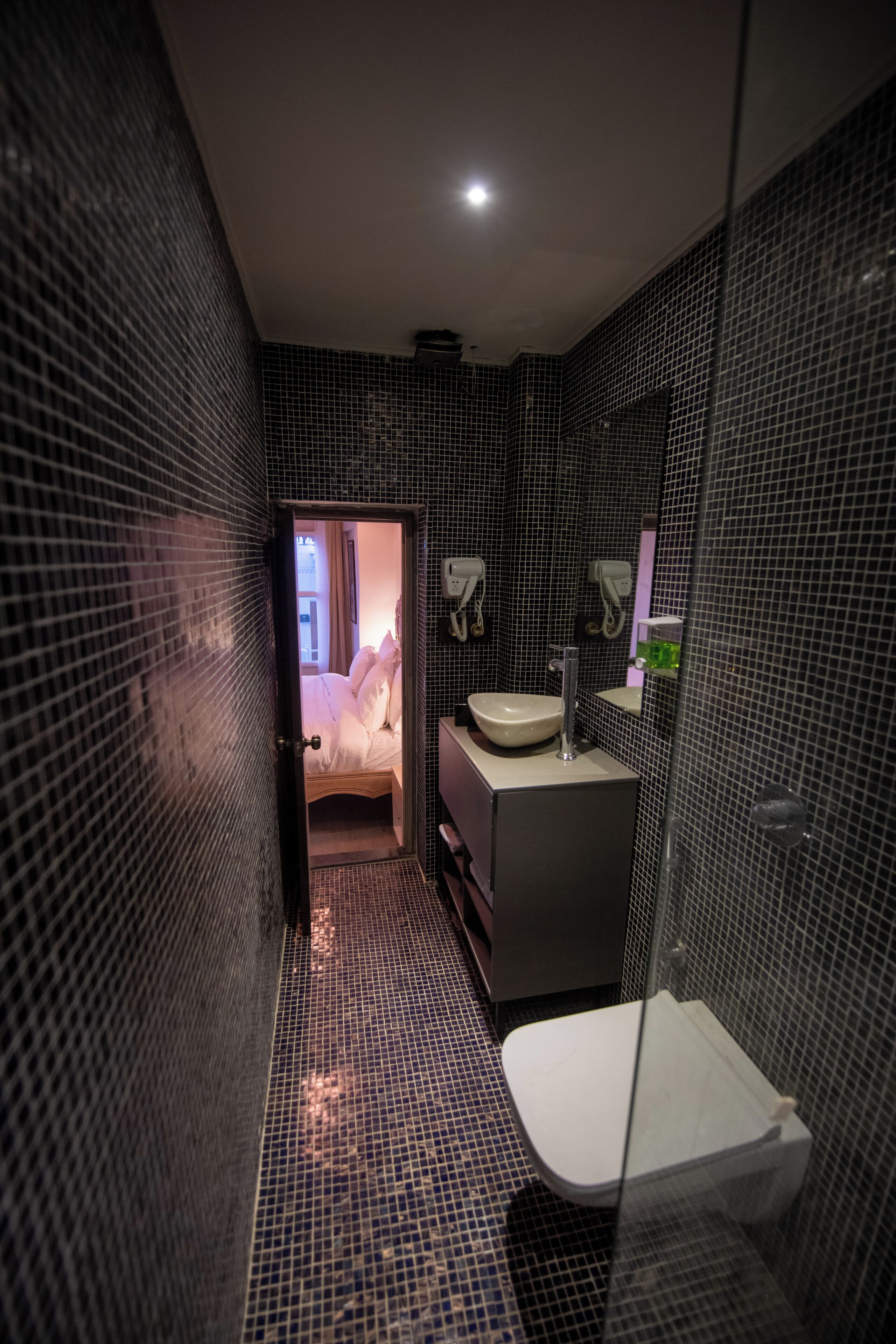 A modern bathroom with a tiled floor, a sink, a toilet, and a bed visible through an open door.