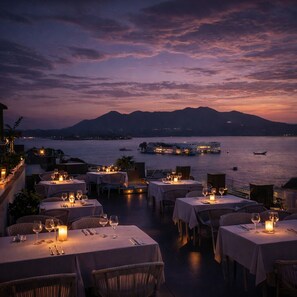 A seaside restaurant with tables set for dining, overlooking a calm body of water and distant mountains. - OTC By 4 Lal Ghat - My Downtown Hotels (Udaipur)