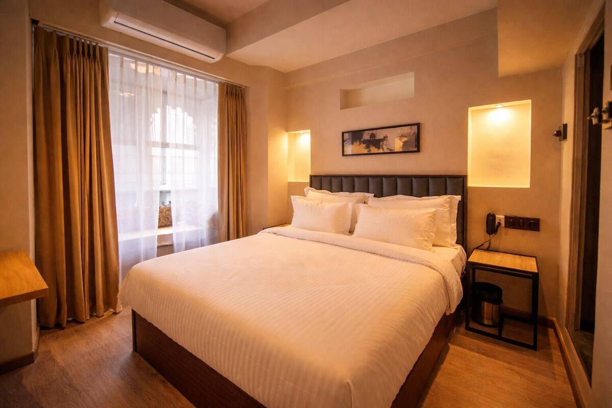 Una habitación de hotel con una cama grande, una mesita de noche, una lámpara de pared y una ventana con cortinas.