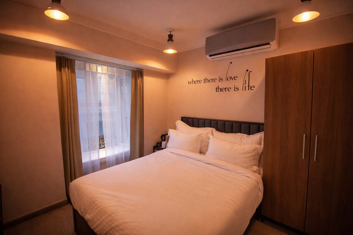 Habitación de hotel con cama, mesita de noche, equipo de aire acondicionado, armario y ventana con cortinas.
