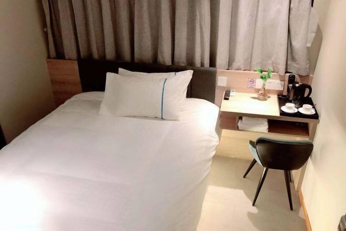 Una cama bien hecha con sábanas blancas, un cabecero de madera, un pequeño escritorio con una silla, una cafetera y una planta en maceta.