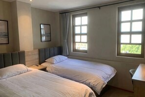 A hotel room with two beds, a desk, a chair, and large windows with curtains.