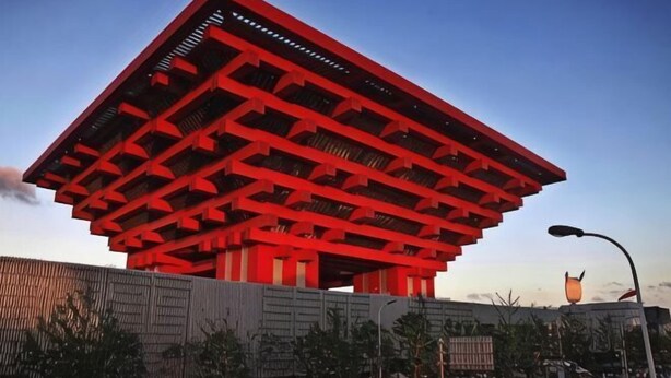 Um edifício grande e vermelho com um design geométrico singular, cercado por uma cerca e postes de luz.