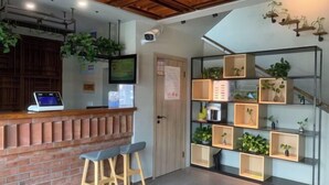 Uma área de recepção com balcão de tijolos, dois banquetes, um computador e uma parede verde com plantas.