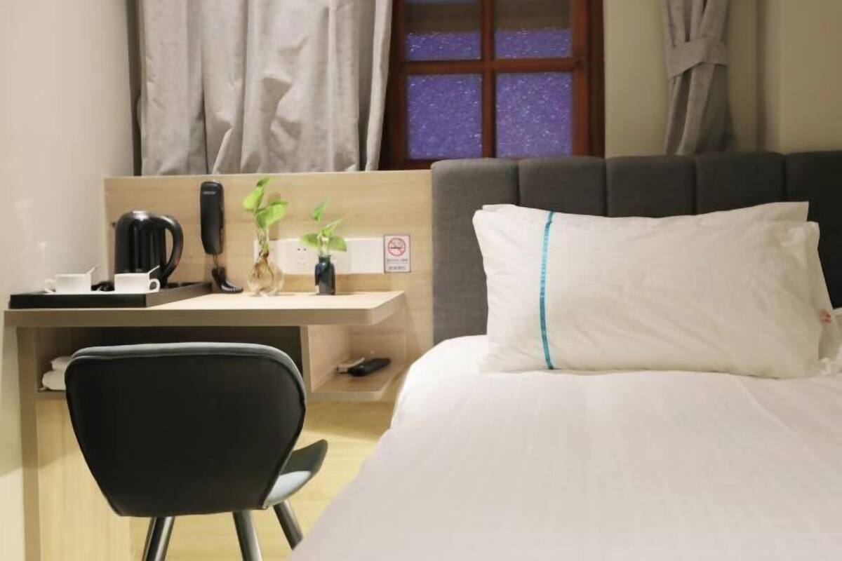 Habitación de hotel con cama, escritorio con computadora, silla, planta y ventana con cortinas.