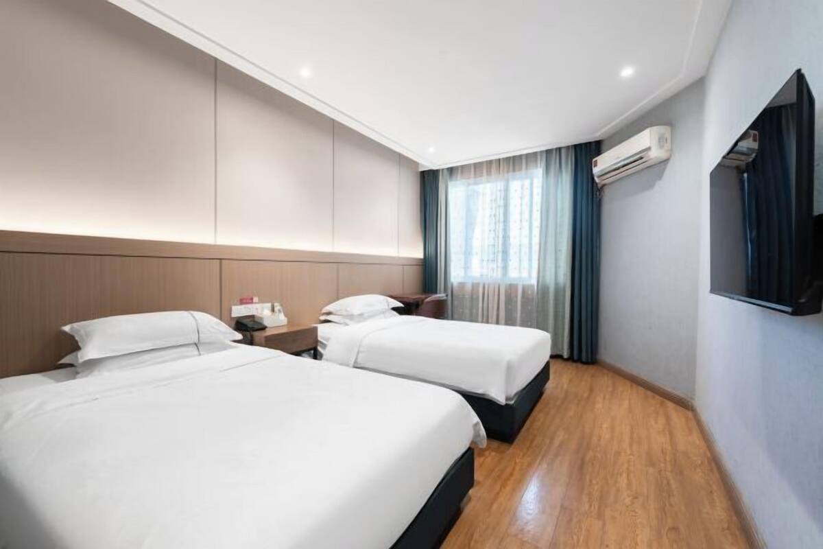 A hotel room with two beds, a wooden floor, a large window with curtains, and an air conditioning unit.