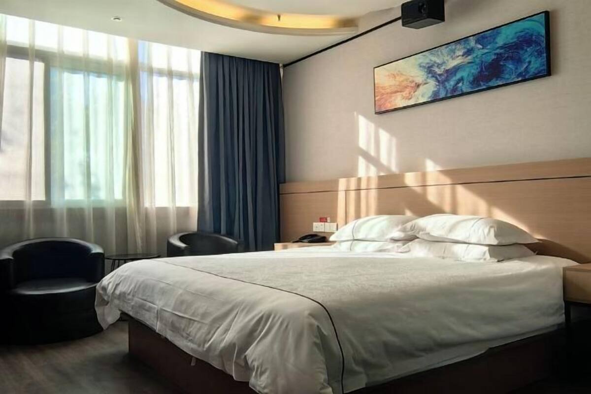 A hotel room with a bed, a chair, a nightstand, a painting, and a window with curtains.