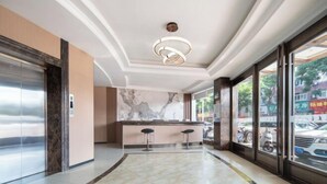 Un vestíbulo de hotel con un mostrador de recepción de mármol, dos taburetes de bar, una lámpara moderna y ventanas amplias que dan a la calle.