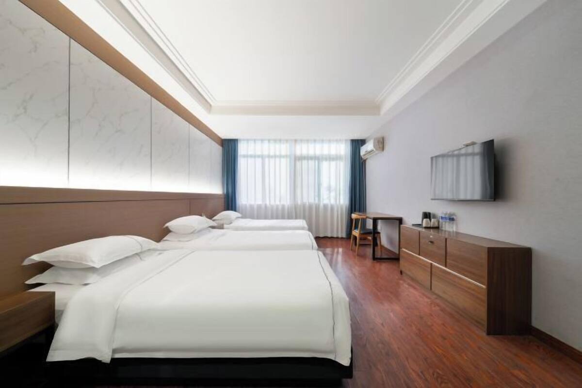 A hotel room with three beds, a wooden headboard, a TV mounted on the wall, a small table, and a cabinet.