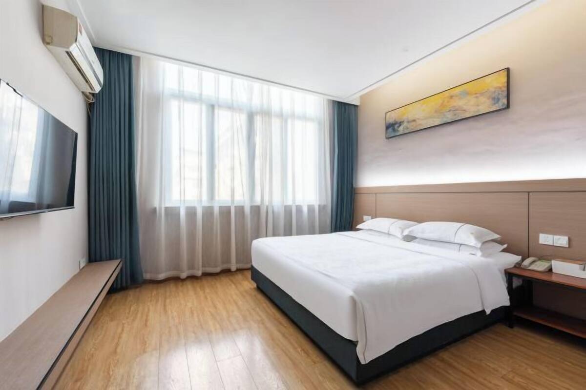 A hotel room with a bed, a TV, a bedside table, a window with curtains, and a painting on the wall.