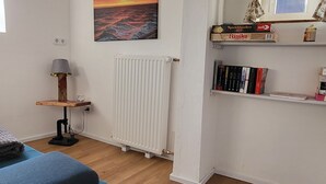 A living room with a blue sofa, a radiator, a painting of a sunset, a lamp, and a bookshelf with books. - Vacation apartment Dörpschipper in the smallest warf village of the Krummhörn (Krummhörn)
