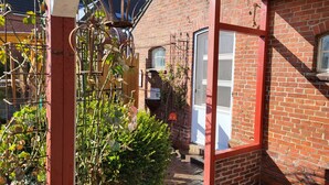 A brick house with a small garden area, a metal trellis with climbing plants, and a white door. - Vacation apartment Dörpschipper in the smallest warf village of the Krummhörn (Krummhörn)
