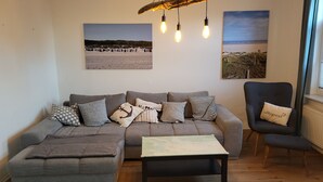 Ein Wohnzimmer mit einem grauen Sofa, dazu passendem Ottomanen und einem schwarzen Sessel. Auf dem Sofa liegen mehrere Kissen, und an der Wand hängen zwei Gemälde.