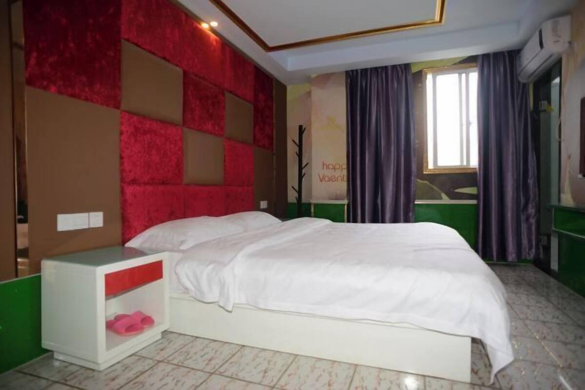 A bedroom with a bed, a nightstand, a red and beige wall panel, a window with curtains, and a sign on the wall.