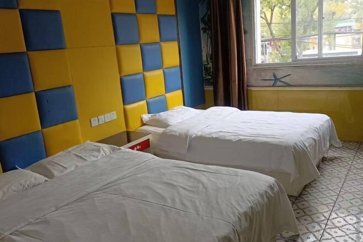 A room with two beds, a window with curtains, a wall with a blue and yellow pattern, and a tiled floor.